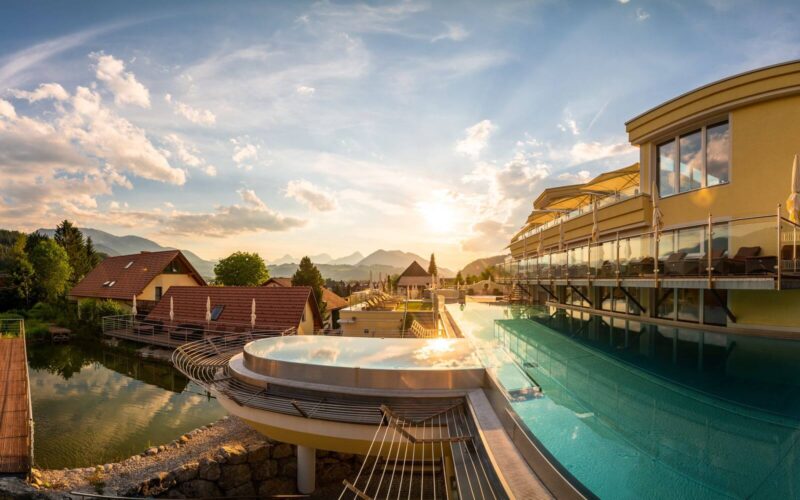 Modernes Gebäude mit einem Infinity-Pool mit Blick auf einen Teich, die Dächer und die fernen Berge bei Sonnenuntergang unter einem teilweise bewölkten Himmel.