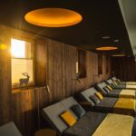Ein Lounge-Bereich mit Holzwänden, mehreren gepolsterten Sesseln mit gelben Decken und Sonnenlicht, das durch ein Fenster fällt.