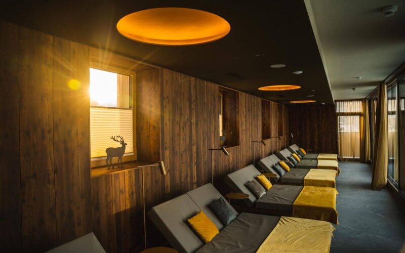 Ein Lounge-Bereich mit Holzwänden, mehreren gepolsterten Sesseln mit gelben Decken und Sonnenlicht, das durch ein Fenster fällt.
