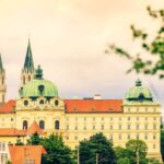 Ein großes historisches Gebäude mit grünen Kuppeln und Zwillingstürmen erhebt sich unter einem wolkenverhangenen Himmel über die umliegenden Bäume und kleineren Gebäude.