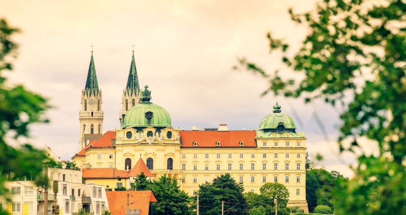 Ein großes historisches Gebäude mit grünen Kuppeln und Zwillingstürmen erhebt sich unter einem wolkenverhangenen Himmel über die umliegenden Bäume und kleineren Gebäude.
