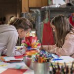 Zwei Mädchen sitzen an einem Tisch, der mit Bastelmaterial bedeckt ist, und zeichnen und machen Kunstprojekte mit Federn, Papier und Stiften in einem Klassenzimmer oder Atelier.