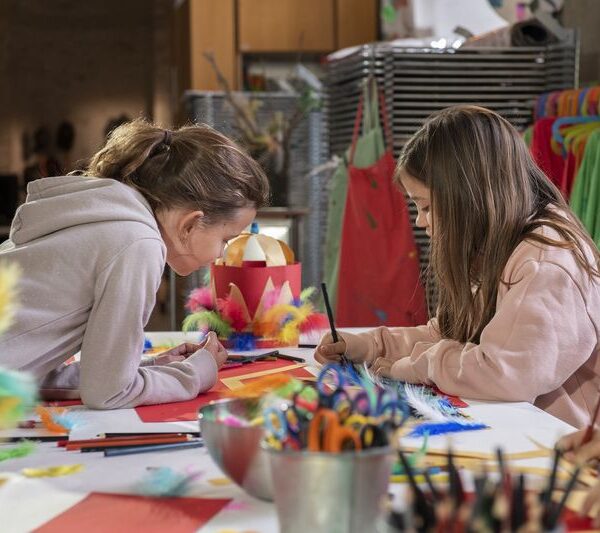 Zwei Mädchen sitzen an einem Tisch, der mit Bastelmaterial bedeckt ist, und zeichnen und machen Kunstprojekte mit Federn, Papier und Stiften in einem Klassenzimmer oder Atelier.