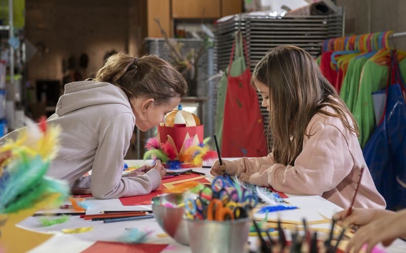 Zwei Mädchen sitzen an einem Tisch, der mit Bastelmaterial bedeckt ist, und zeichnen und machen Kunstprojekte mit Federn, Papier und Stiften in einem Klassenzimmer oder Atelier.
