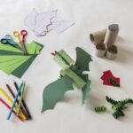 Ein Papierdrache aus grünem Papier, Pappröhren und Kulleraugen, umgeben von Scheren, Buntstiften und weiteren Bastelmaterialien auf einer weißen Fläche.