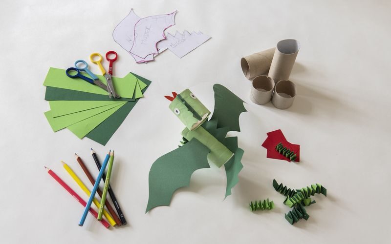 Ein Papierdrache aus grünem Papier, Pappröhren und Kulleraugen, umgeben von Scheren, Buntstiften und weiteren Bastelmaterialien auf einer weißen Fläche.