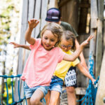 Drei Kinder überqueren eine Seilbrücke im Freien vor einer Holzkonstruktion, wobei das Mädchen im rosa Hemd voran geht und mit ausgestreckten Armen balanciert.