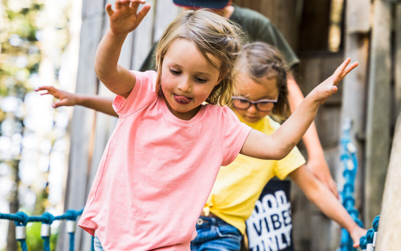 Drei Kinder überqueren eine Seilbrücke im Freien vor einer Holzkonstruktion, wobei das Mädchen im rosa Hemd voran geht und mit ausgestreckten Armen balanciert.