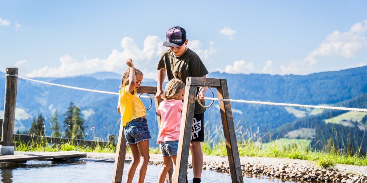 Drei Kinder stehen auf einer hölzernen Plattform über seichtem Wasser und halten sich an Seilen fest. Im Hintergrund sind Hügel und ein blauer Himmel zu sehen.