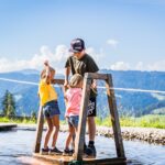 Drei Kinder stehen auf einer hölzernen Plattform über seichtem Wasser und halten sich an Seilen fest. Im Hintergrund sind Hügel und ein blauer Himmel zu sehen.