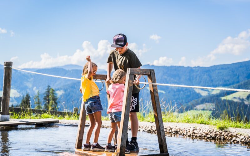Drei Kinder stehen auf einer hölzernen Plattform über seichtem Wasser und halten sich an Seilen fest. Im Hintergrund sind Hügel und ein blauer Himmel zu sehen.