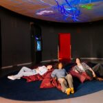 Drei Personen liegen auf Sitzsäcken in einem dunklen Raum und blicken auf eine Decke mit blauen und grünen Leuchtmustern.