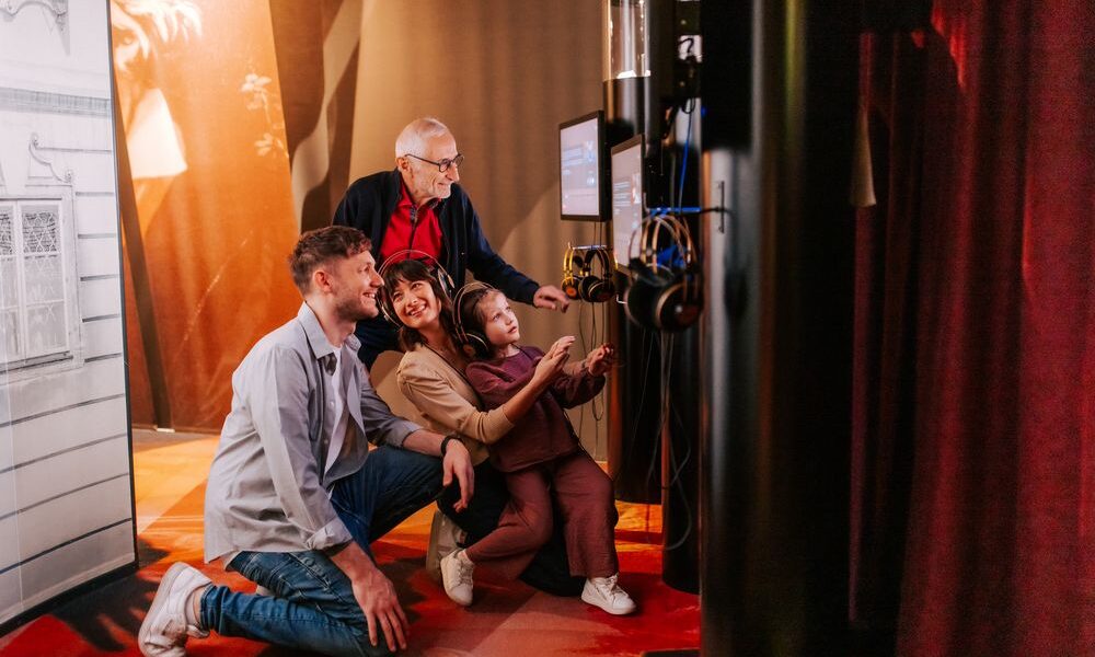 Vier Personen unterschiedlichen Alters interagieren mit einem Touchscreen und Kopfhörern in einer schwach beleuchteten Ausstellung oder einem Museum.