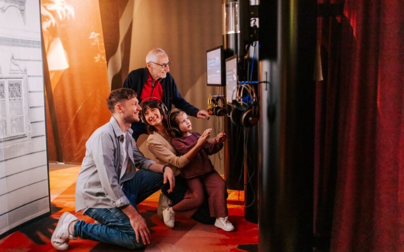 Vier Personen unterschiedlichen Alters interagieren mit einem Touchscreen und Kopfhörern in einer schwach beleuchteten Ausstellung oder einem Museum.