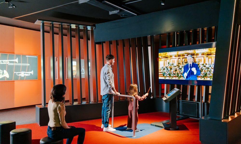 Ein Mann und ein Kind interagieren mit einem Touchscreen in einem modernen Ausstellungsraum, während eine Frau in der Nähe sitzt; ein großer Bildschirm zeigt eine Person, die in einer formellen Umgebung spricht.