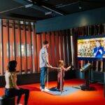 Ein Mann und ein Kind interagieren mit einem Touchscreen in einem modernen Ausstellungsraum, während eine Frau in der Nähe sitzt; ein großer Bildschirm zeigt eine Person, die in einer formellen Umgebung spricht.