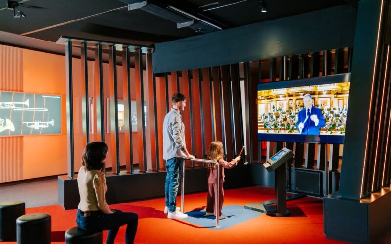 Ein Mann und ein Kind interagieren mit einem Touchscreen in einem modernen Ausstellungsraum, während eine Frau in der Nähe sitzt; ein großer Bildschirm zeigt eine Person, die in einer formellen Umgebung spricht.