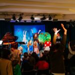 Kinder und Erwachsene erheben ihre Arme während einer lebhaften Aufführung auf der Bühne mit einem Pianisten, farbenfrohen Kostümen und einer skurrilen Kulisse mit Tier- und Naturelementen.