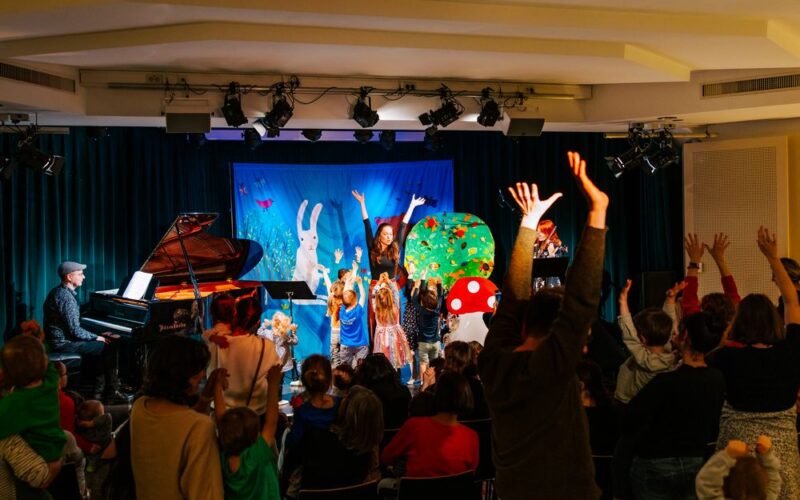 Kinder und Erwachsene erheben ihre Arme während einer lebhaften Aufführung auf der Bühne mit einem Pianisten, farbenfrohen Kostümen und einer skurrilen Kulisse mit Tier- und Naturelementen.