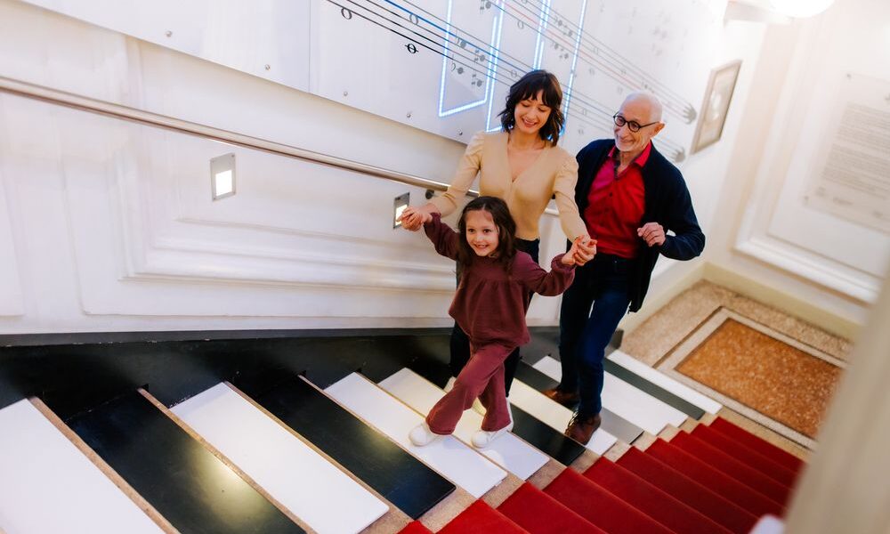 Drei Personen - ein älterer Mann, eine Frau und ein junges Mädchen - gehen lächelnd und händchenhaltend eine Treppe hinauf, die wie Klaviertasten aussieht.