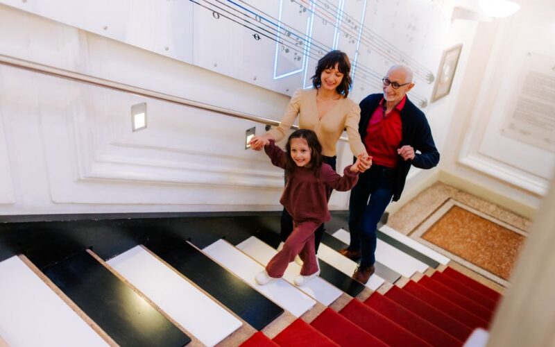 Drei Personen - ein älterer Mann, eine Frau und ein junges Mädchen - gehen lächelnd und händchenhaltend eine Treppe hinauf, die wie Klaviertasten aussieht.