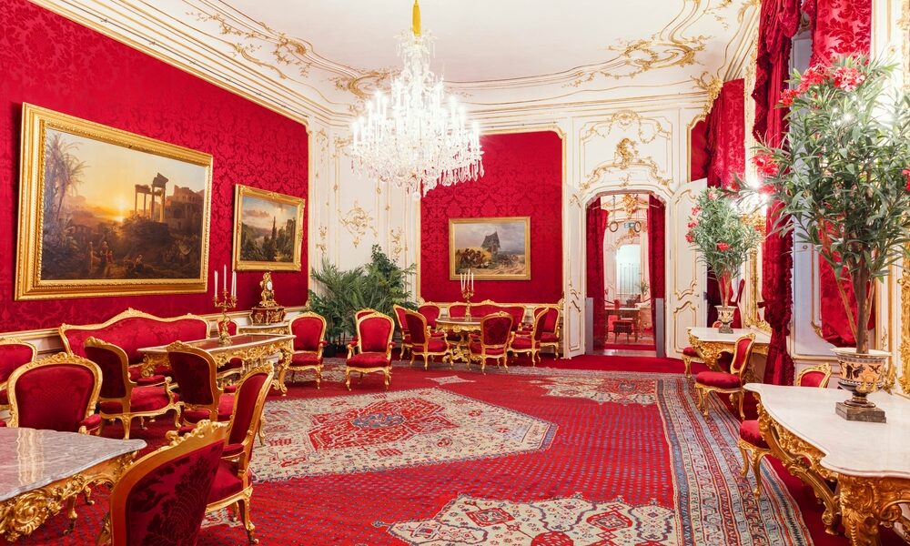 Prunkvoller Raum mit roten Wänden, goldverzierten Möbeln und Spiegeln, einem Kristalllüster, großen Gemälden, verzierten Teppichen und hohen Pflanzen.