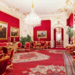 Prunkvoller Raum mit roten Wänden, goldverzierten Möbeln und Spiegeln, einem Kristalllüster, großen Gemälden, verzierten Teppichen und hohen Pflanzen.
