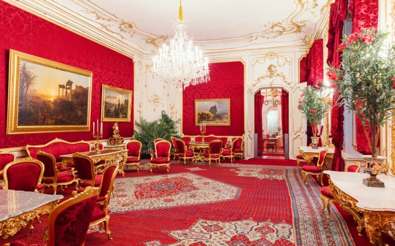 Prunkvoller Raum mit roten Wänden, goldverzierten Möbeln und Spiegeln, einem Kristalllüster, großen Gemälden, verzierten Teppichen und hohen Pflanzen.