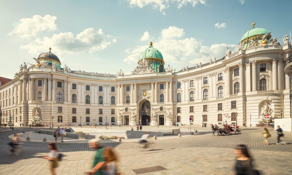 Historisches weißes Barockgebäude mit grünen Kuppeln, verzierten Details und einem gewölbten Eingang, vor dem die Menschen auf dem kopfsteingepflasterten Platz spazieren gehen.