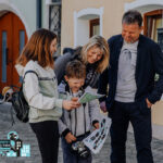 Eine vierköpfige Familie steht im Freien auf einer Kopfsteinpflasterstraße, schaut sich gemeinsam Karten und Papiere an, lächelt und ist leger gekleidet.