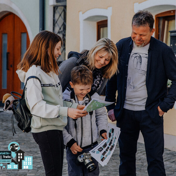 Eine vierköpfige Familie steht im Freien auf einer Kopfsteinpflasterstraße, schaut sich gemeinsam Karten und Papiere an, lächelt und ist leger gekleidet.