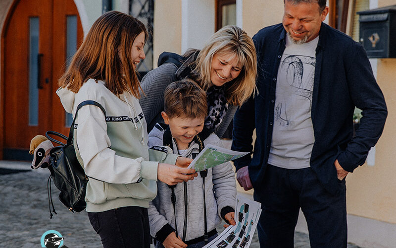 Eine vierköpfige Familie steht im Freien auf einer Kopfsteinpflasterstraße, schaut sich gemeinsam Karten und Papiere an, lächelt und ist leger gekleidet.