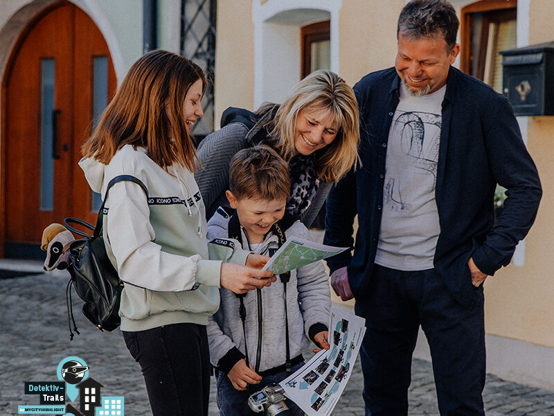 Eine vierköpfige Familie steht im Freien auf einer Kopfsteinpflasterstraße, schaut sich gemeinsam Karten und Papiere an, lächelt und ist leger gekleidet.