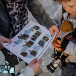 Drei Personen, darunter ein Kind mit einem Spielzeugdachs und einer Kamera, halten ein gedrucktes Arbeitsblatt mit Fotos und Text in der Hand und untersuchen es.
