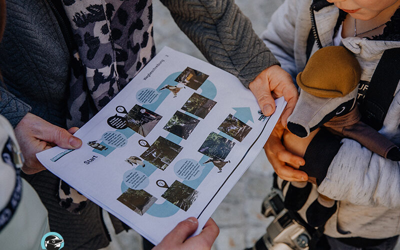 Drei Personen, darunter ein Kind mit einem Spielzeugdachs und einer Kamera, halten ein gedrucktes Arbeitsblatt mit Fotos und Text in der Hand und untersuchen es.