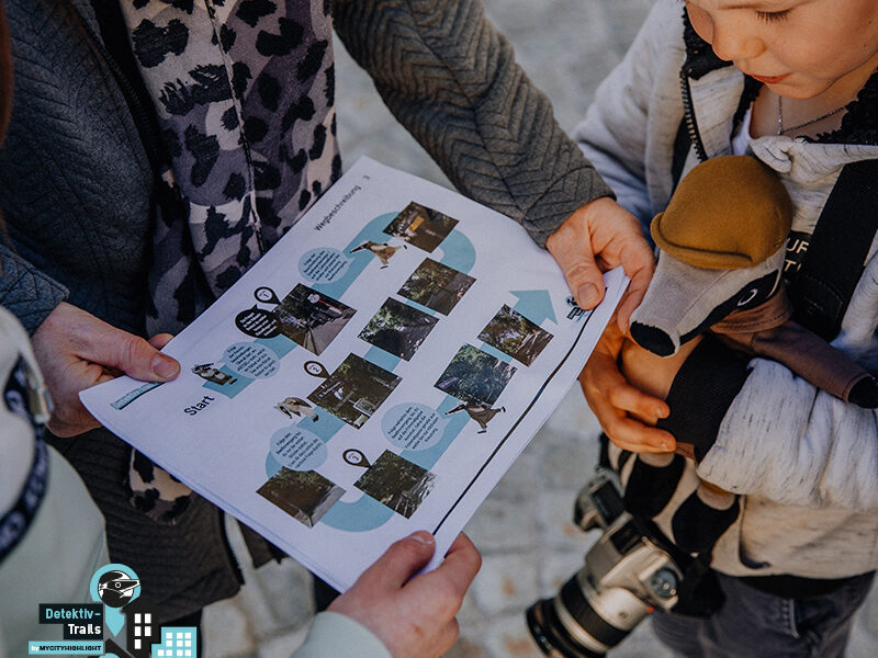 Drei Personen, darunter ein Kind mit einem Spielzeugdachs und einer Kamera, halten ein gedrucktes Arbeitsblatt mit Fotos und Text in der Hand und untersuchen es.