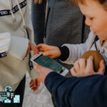 Drei Kinder stehen zusammen, eines hält ein Smartphone, ein anderes zeigt auf den Bildschirm. Auf dem Bildschirm wird Text angezeigt. Ein Kind hält ein rundes braunes Objekt.