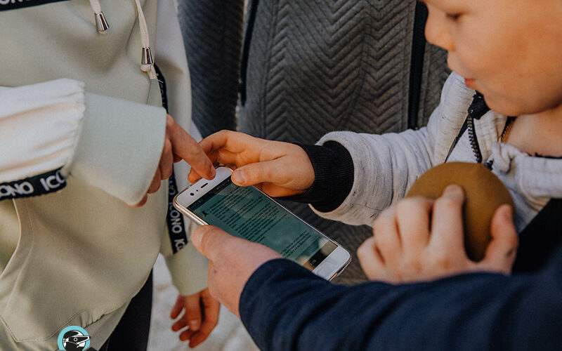 Drei Kinder stehen zusammen, eines hält ein Smartphone, ein anderes zeigt auf den Bildschirm. Auf dem Bildschirm wird Text angezeigt. Ein Kind hält ein rundes braunes Objekt.