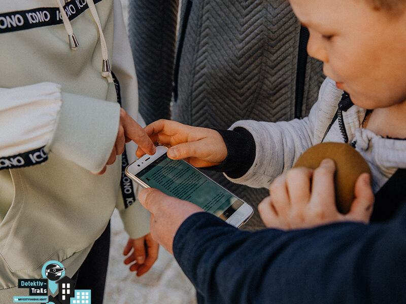 Drei Kinder stehen zusammen, eines hält ein Smartphone, ein anderes zeigt auf den Bildschirm. Auf dem Bildschirm wird Text angezeigt. Ein Kind hält ein rundes braunes Objekt.