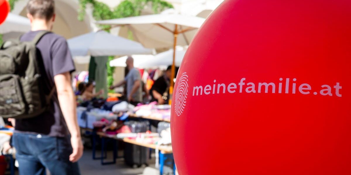 Im Fokus steht ein roter Ballon mit der Aufschrift „meinefamilie.at“; im unscharfen Hintergrund sind Menschen und Marktstände im Freien zu sehen.