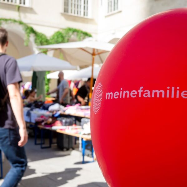Im Fokus steht ein roter Ballon mit der Aufschrift „meinefamilie.at“; im unscharfen Hintergrund sind Menschen und Marktstände im Freien zu sehen.