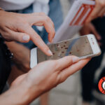 Nahaufnahme einer Person, die ein Smartphone mit einer geöffneten Karten-App auf dem Bildschirm benutzt; im Hintergrund sind andere Personen und ein Krimi Trails-Logo zu sehen.