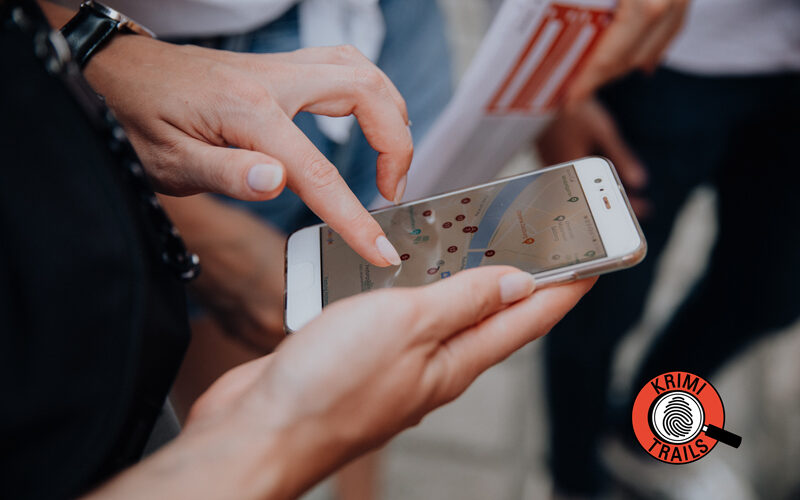 Nahaufnahme einer Person, die ein Smartphone mit einer geöffneten Karten-App auf dem Bildschirm benutzt; im Hintergrund sind andere Personen und ein Krimi Trails-Logo zu sehen.