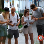 Vier Personen stehen auf einem Gehweg, schauen auf Papiere und ein Telefon und scheinen zusammenzuarbeiten oder etwas zu lösen; in der Ecke ist ein "Krimi Trails"-Logo zu sehen.