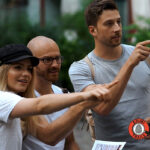 Drei Erwachsene stehen im Freien und zeigen alle interessiert in die gleiche Richtung. Einer hält eine Zeitung in der Hand, ein anderer trägt einen Hut. In der rechten unteren Ecke befindet sich ein "Krimi Trails"-Logo.