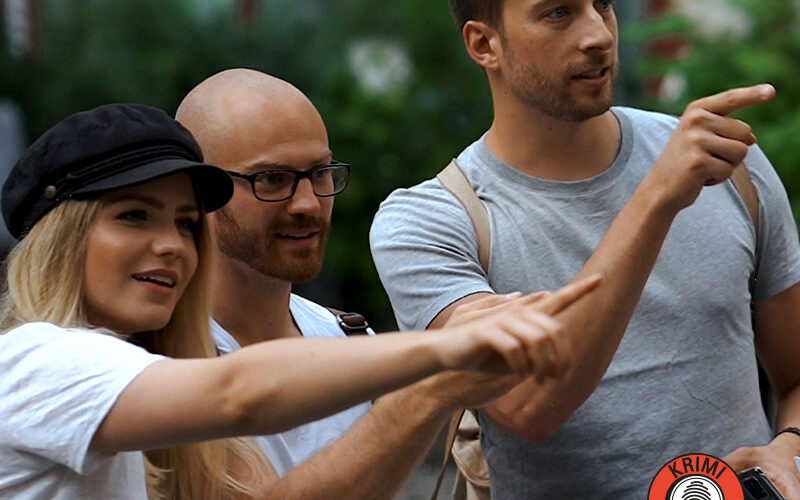 Drei Erwachsene stehen im Freien und zeigen alle interessiert in die gleiche Richtung. Einer hält eine Zeitung in der Hand, ein anderer trägt einen Hut. In der rechten unteren Ecke befindet sich ein "Krimi Trails"-Logo.