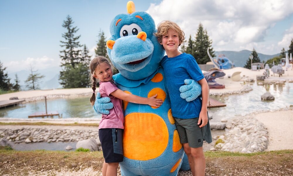 Zwei Kinder stehen im Freien, lächeln und umarmen eine Person in einem blau-orangefarbenen Dinosaurierkostüm, im Hintergrund sind ein Teich und Bäume zu sehen.