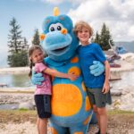 Zwei Kinder stehen im Freien, lächeln und umarmen eine Person in einem blau-orangefarbenen Dinosaurierkostüm, im Hintergrund sind ein Teich und Bäume zu sehen.