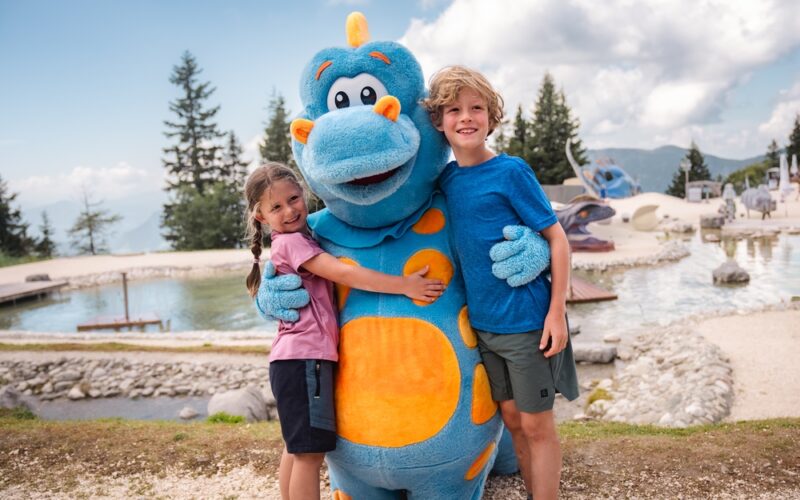 Zwei Kinder stehen im Freien, lächeln und umarmen eine Person in einem blau-orangefarbenen Dinosaurierkostüm, im Hintergrund sind ein Teich und Bäume zu sehen.