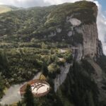 Eine runde hölzerne Aussichtsplattform überblickt einen steilen, bewaldeten Berghang mit einer felsigen Steilwand und einem wolkenverhangenen Himmel im Hintergrund.